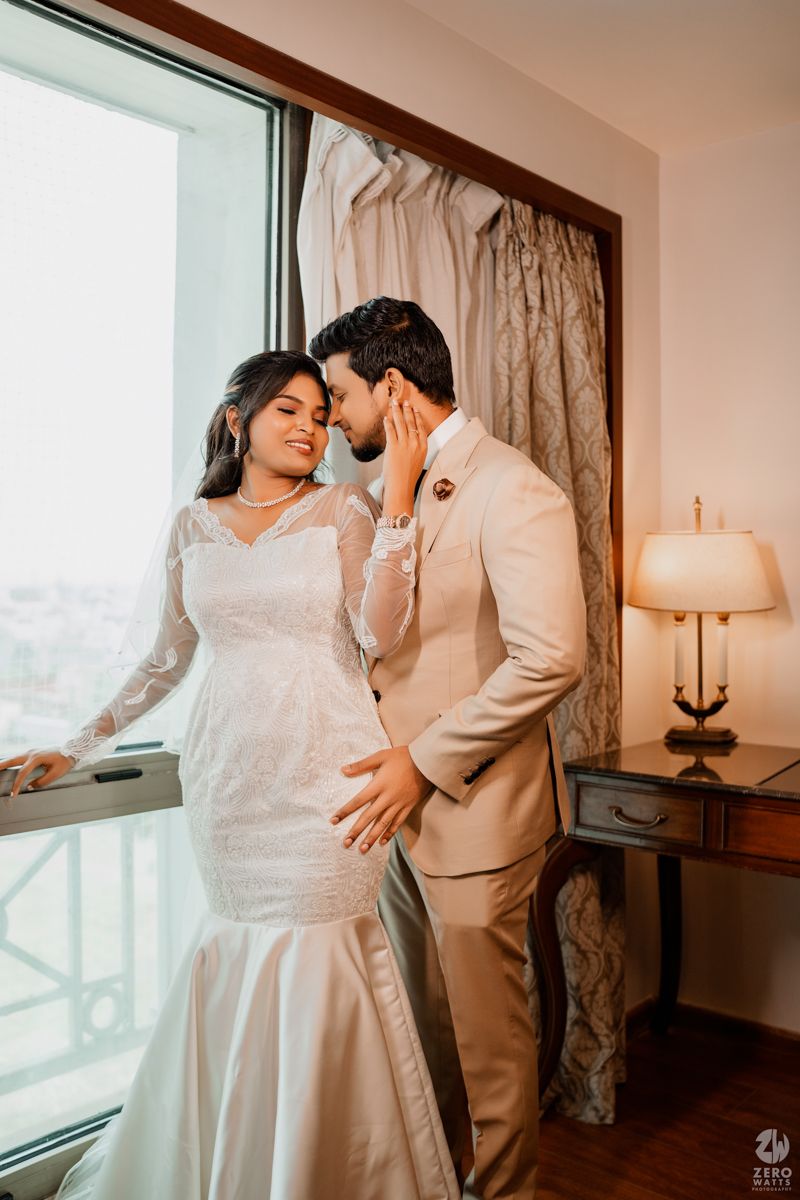 Romantic wedding couple in white and beige outfits posing near a window in an intimate setting.