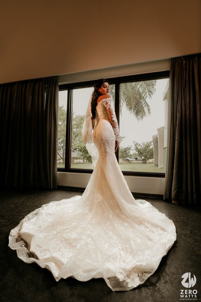 A stunning lace mermaid wedding dress with a long cathedral train fills the frame in a moody indoor setting.