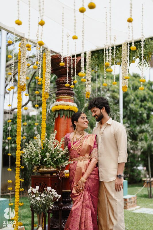Couple in traditional wedding attire standing under marigold and floral decorations at outdoor venue.