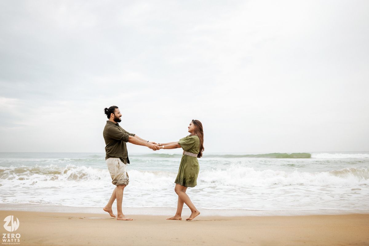 Two people in casual attire dancing together on a sandy beach with overcast skies.