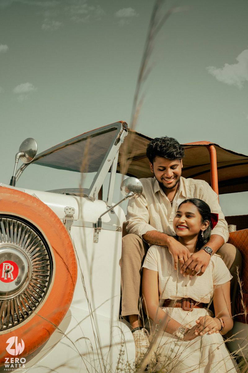 A couple in light colored outfits share a romantic moment beside a vintage white and orange car under a cloudy sky.