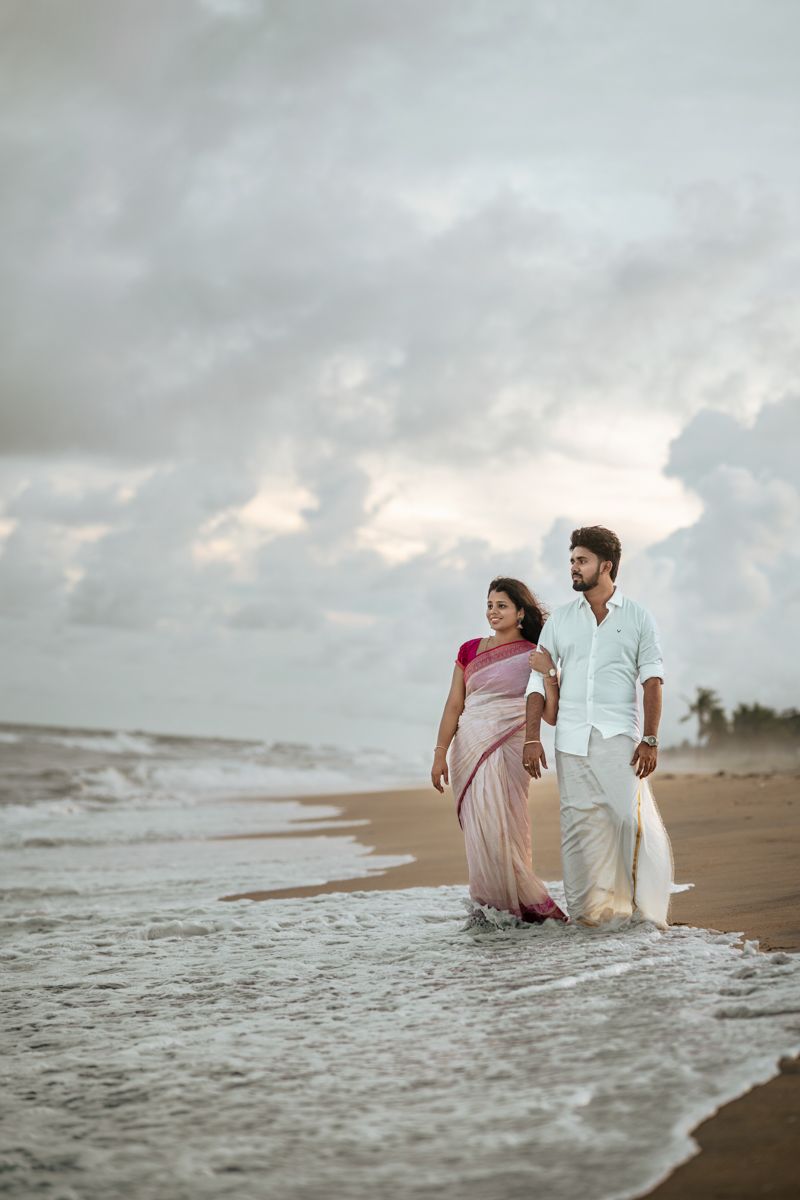 Couple in white and pink attire playfully walking through ocean waves at sunset.
