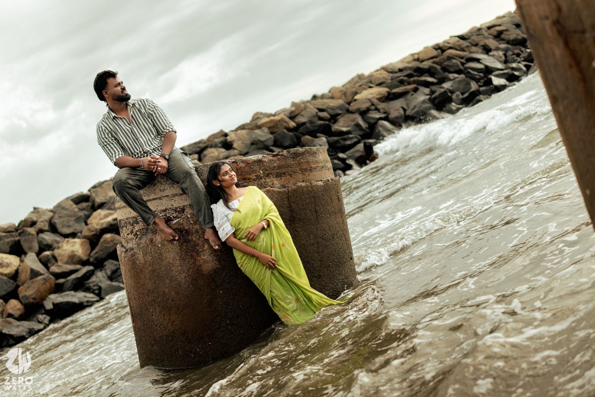 Couple sits on concrete pillars by rocky shoreline during overcast day.