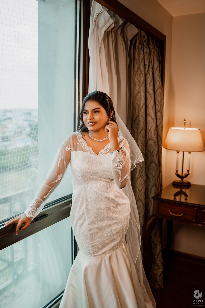 A bride in an elegant white lace mermaid wedding gown and veil poses by a window with a table lamp nearby.