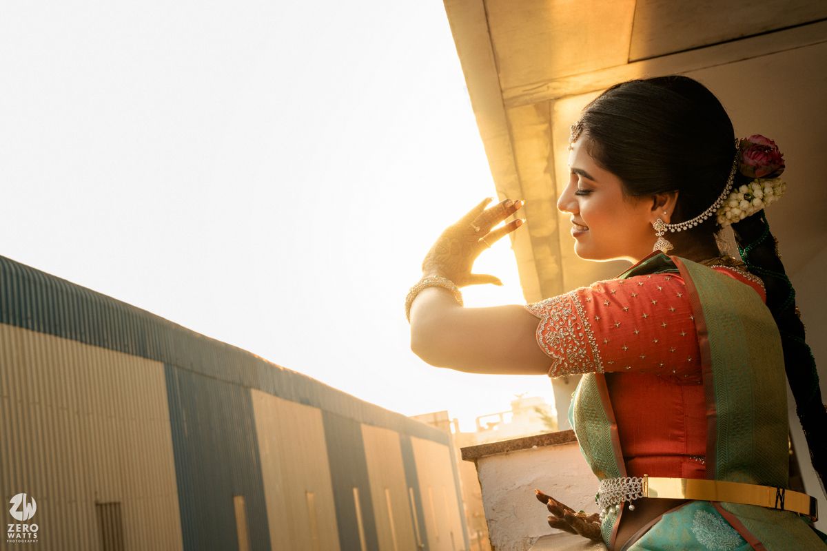 South Indian bride in teal saree and orange blouse poses gracefully in golden sunlight during bridal photoshoot.