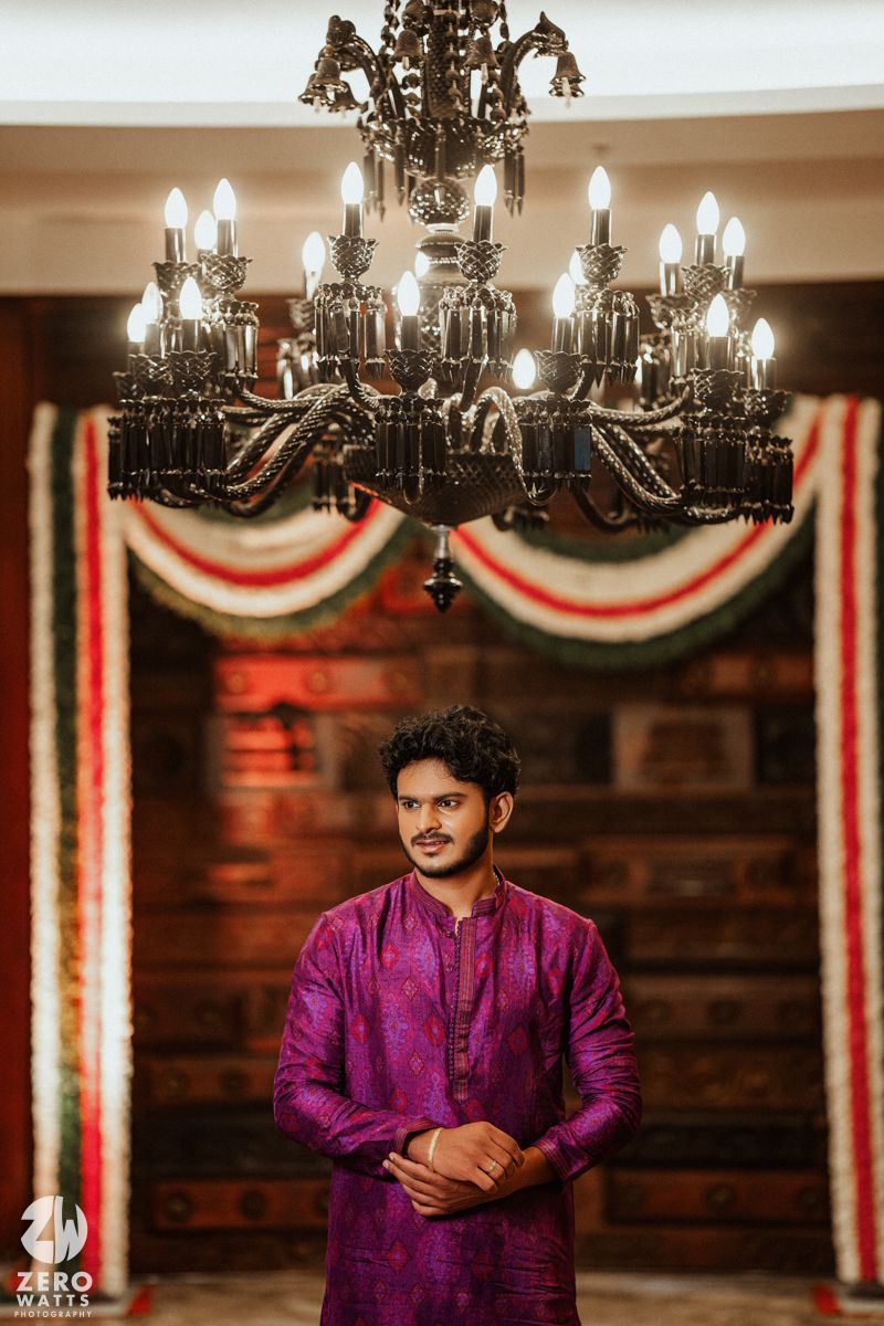 Man in purple kurta stands beneath ornate chandelier with festive red, white, green garland decor backdrop.