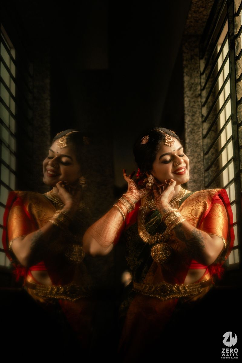 South Indian bride in red silk saree with gold jewelry smiling, reflected in a mirror creating a stunning double portrait.