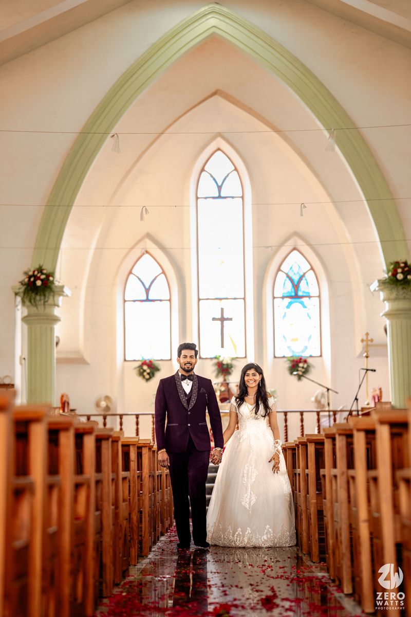 Newlywed couple walking down church aisle lined with wooden pews and rose petals after ceremony.