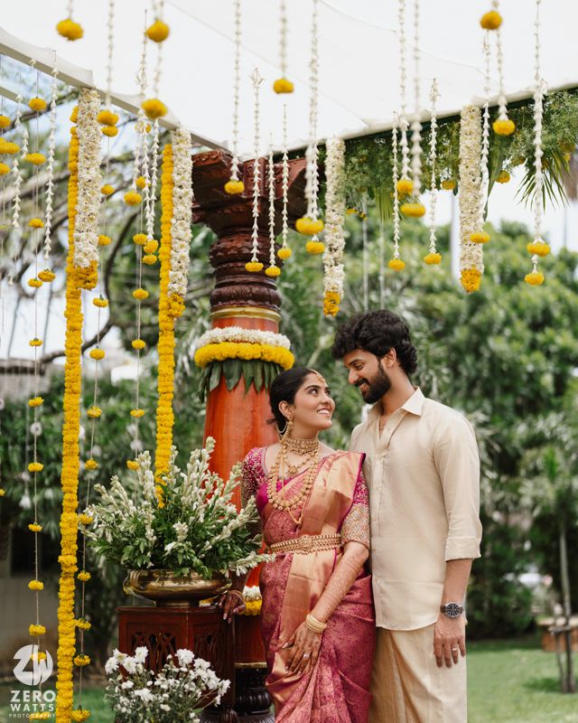 Couple in traditional wedding attire pose under hanging marigold decorations in garden setting.