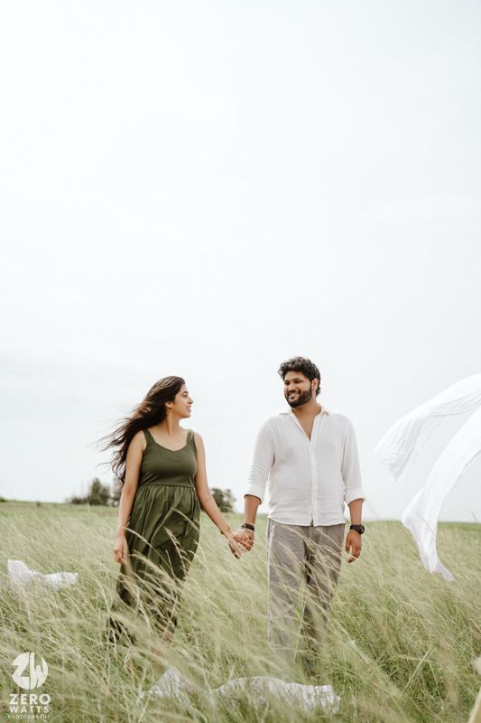 A couple holding hands while walking through tall grass in a meadow wearing casual white and olive green outfits.