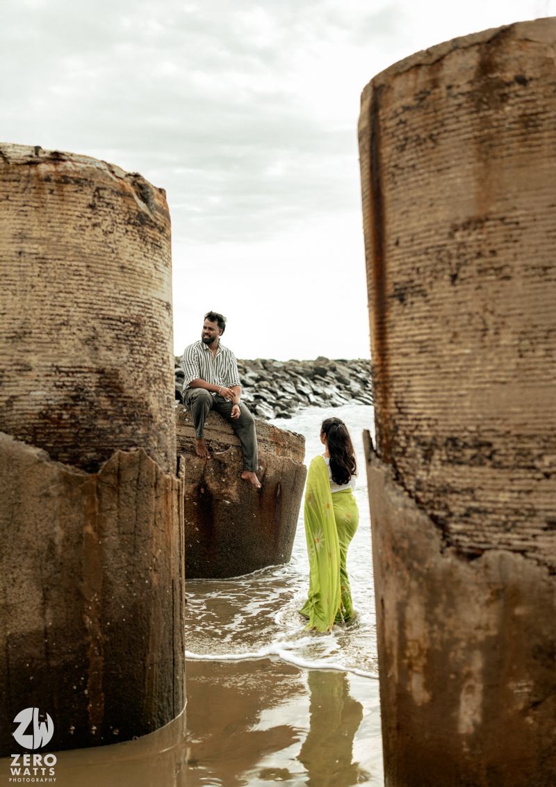 A couple poses artistically between large concrete structures at a beach with moody skies.