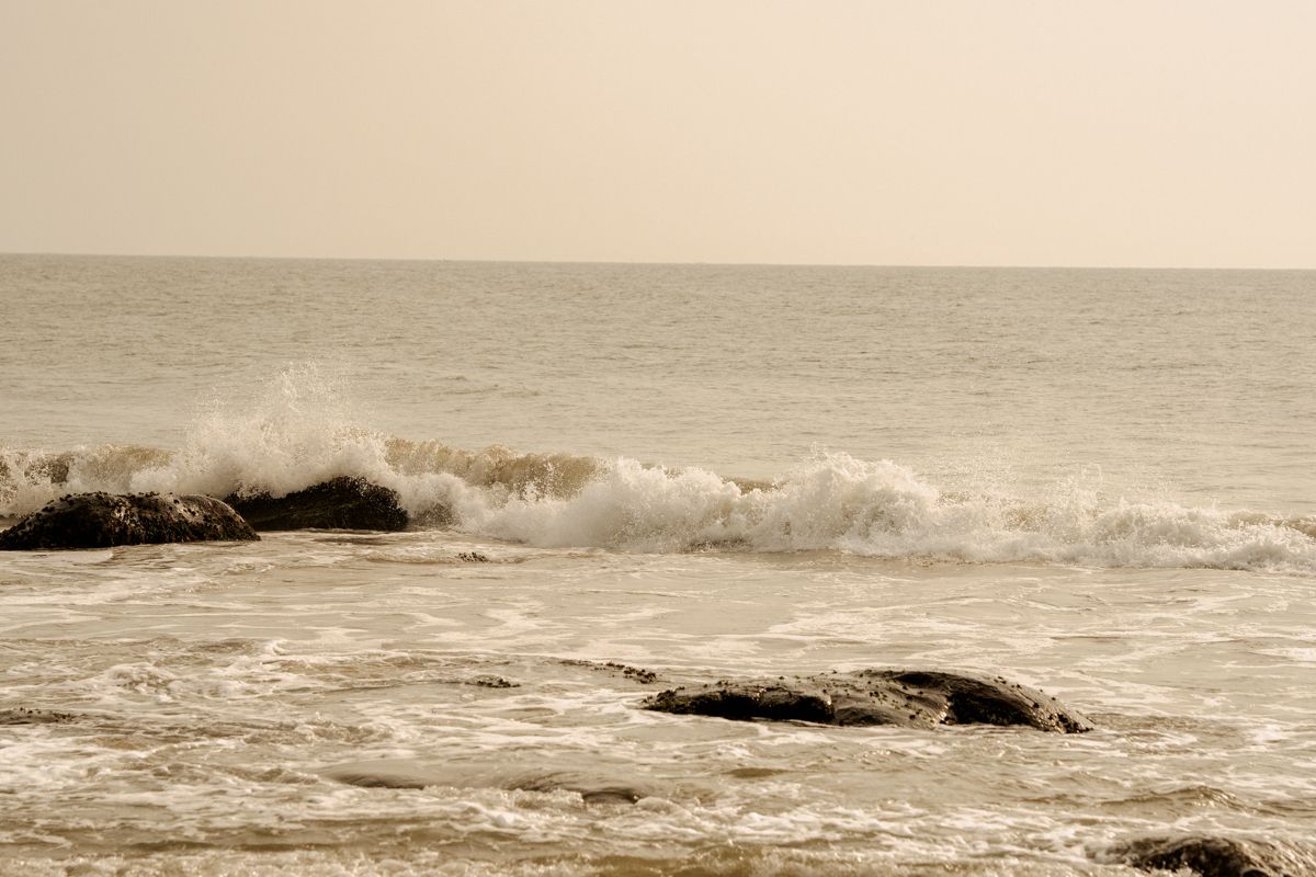 Panoramic seascape showing crashing ocean waves against rocky outcrops in muted sepia tones.