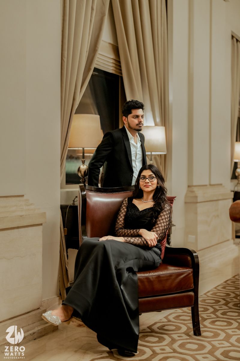 Couple in formal attire poses on leather sofa in elegant room with cream curtains.