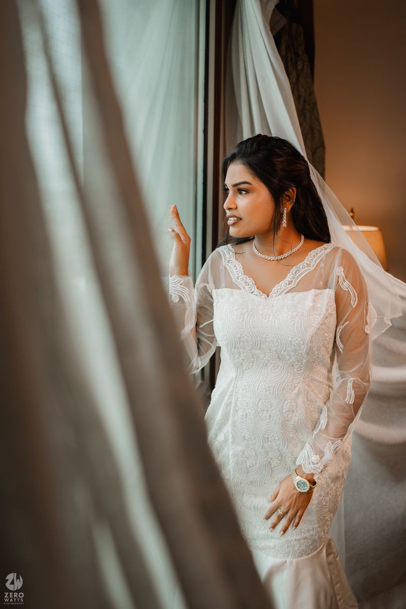 Soft light streams through curtains as a bride in a delicate lace gown stands in a dreamy setting.