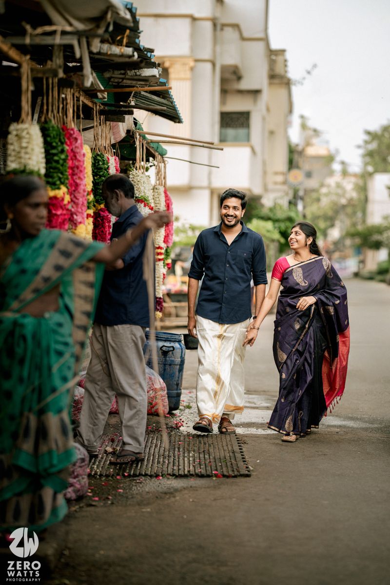 A couple in traditional Indian attire walking and interacting on a street with buildings in the background.
