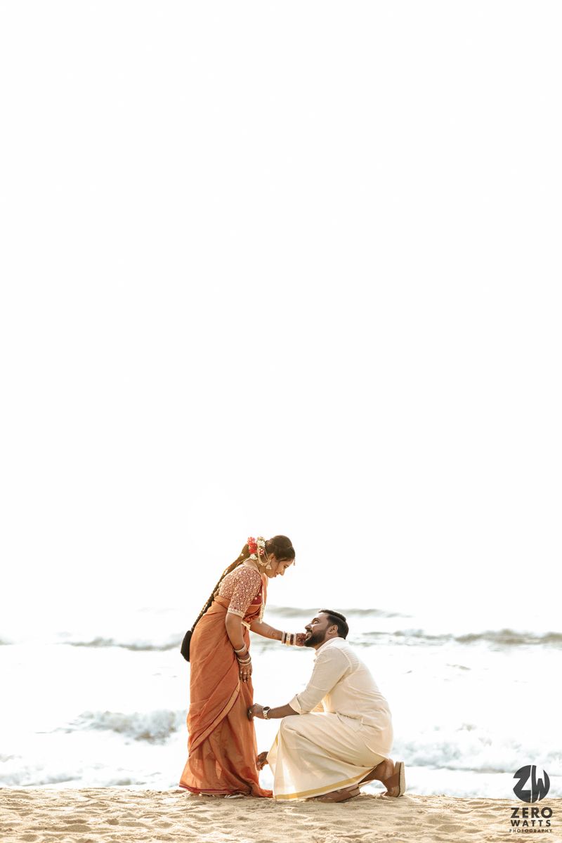 A couple in casual attire sits together on a sandy beach with ocean waves in the background.