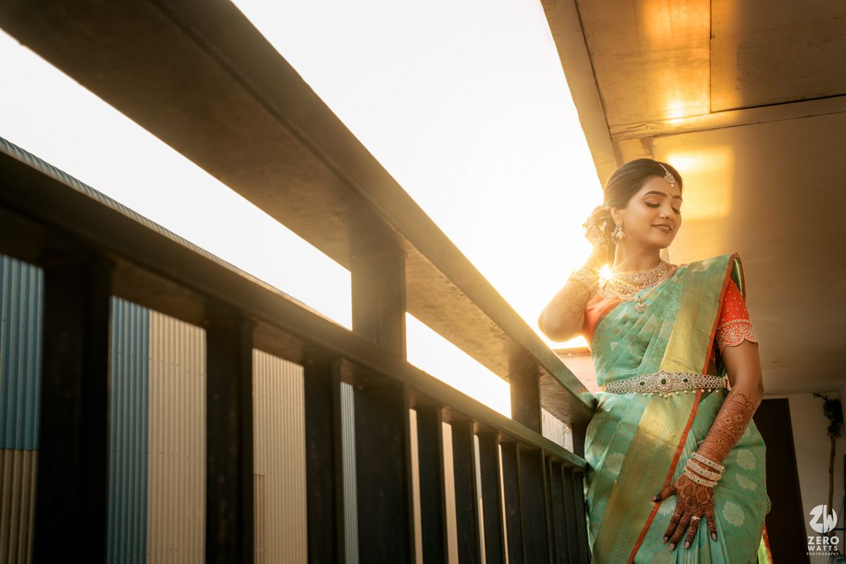 Bride in teal silk saree with gold border stands on sunlit balcony, glowing in warm backlight.