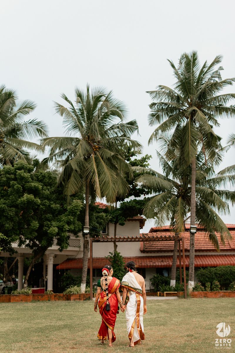 Indian bride in red saree and groom in white dhoti walking on lawn with tall palm trees and traditional venue backdrop.