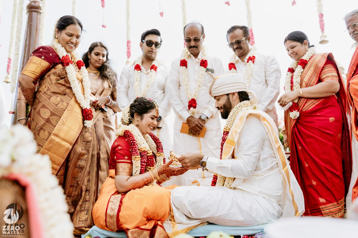Traditional South Indian wedding ceremony with bride in red saree and groom in white sherwani exchanging rituals.