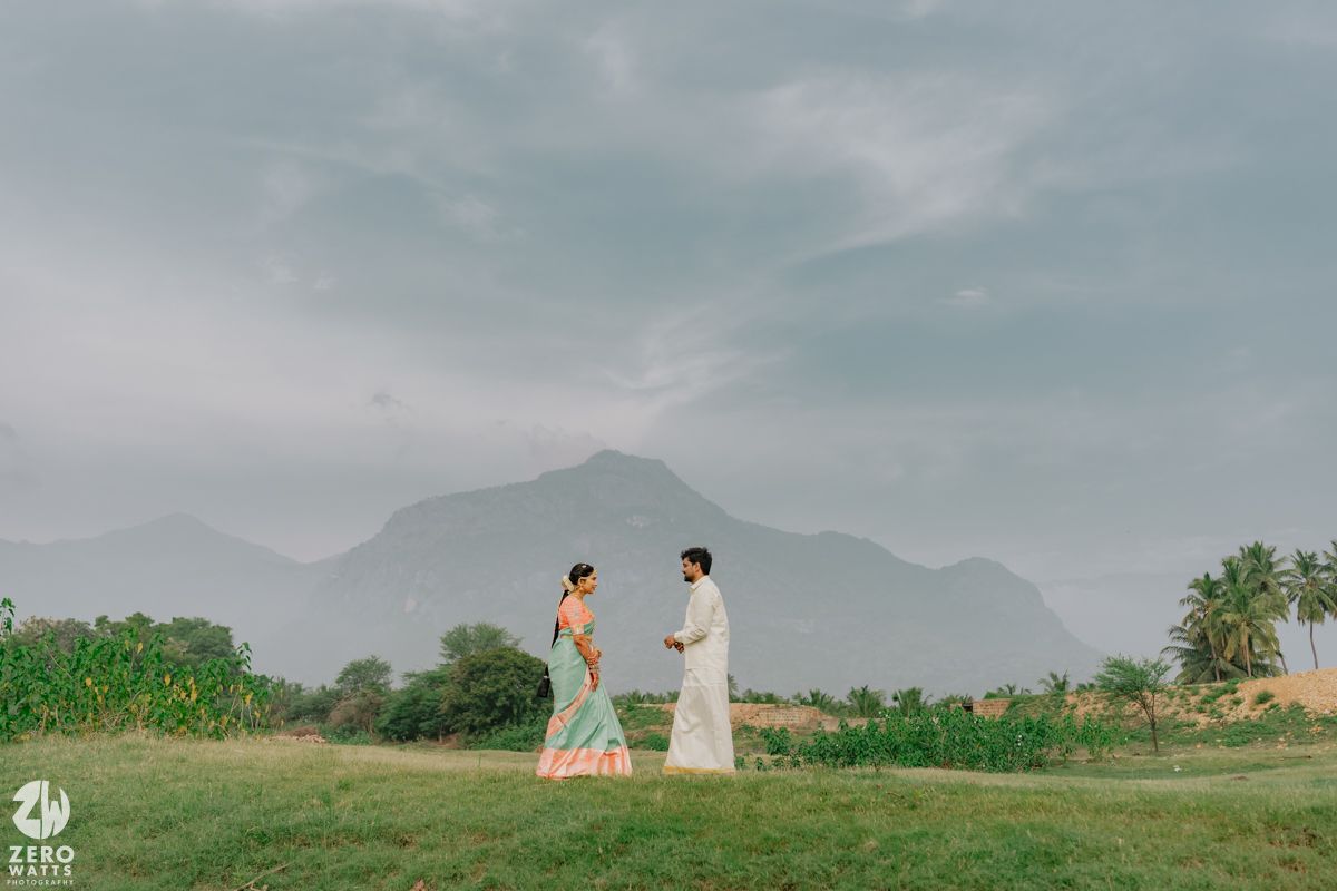 Traditional Indian wedding couple facing each other on green field with mountains.