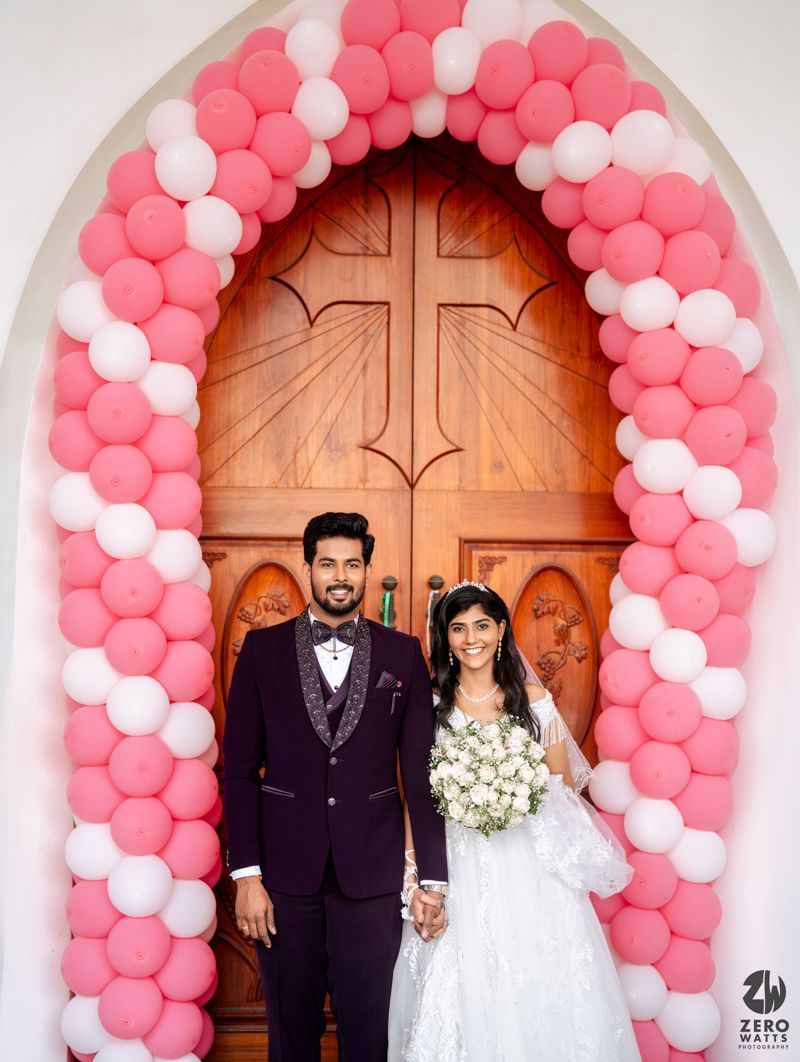 Couple poses under pink and white balloon arch at church entrance with wooden cross door.