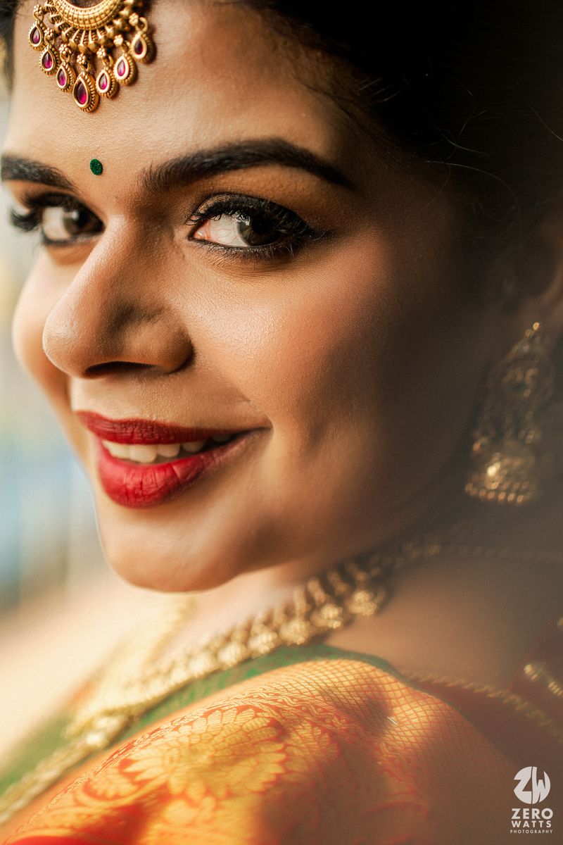South Indian bride smiling in green Kanjivaram saree with gold maang tikka, bindi, and red lips.