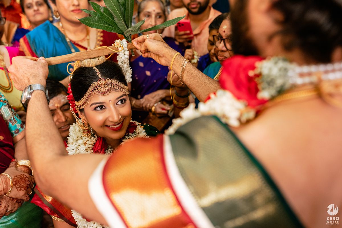Bride in traditional South Indian wedding attire smiles during a Hindu marriage ceremony ritual with garlands.