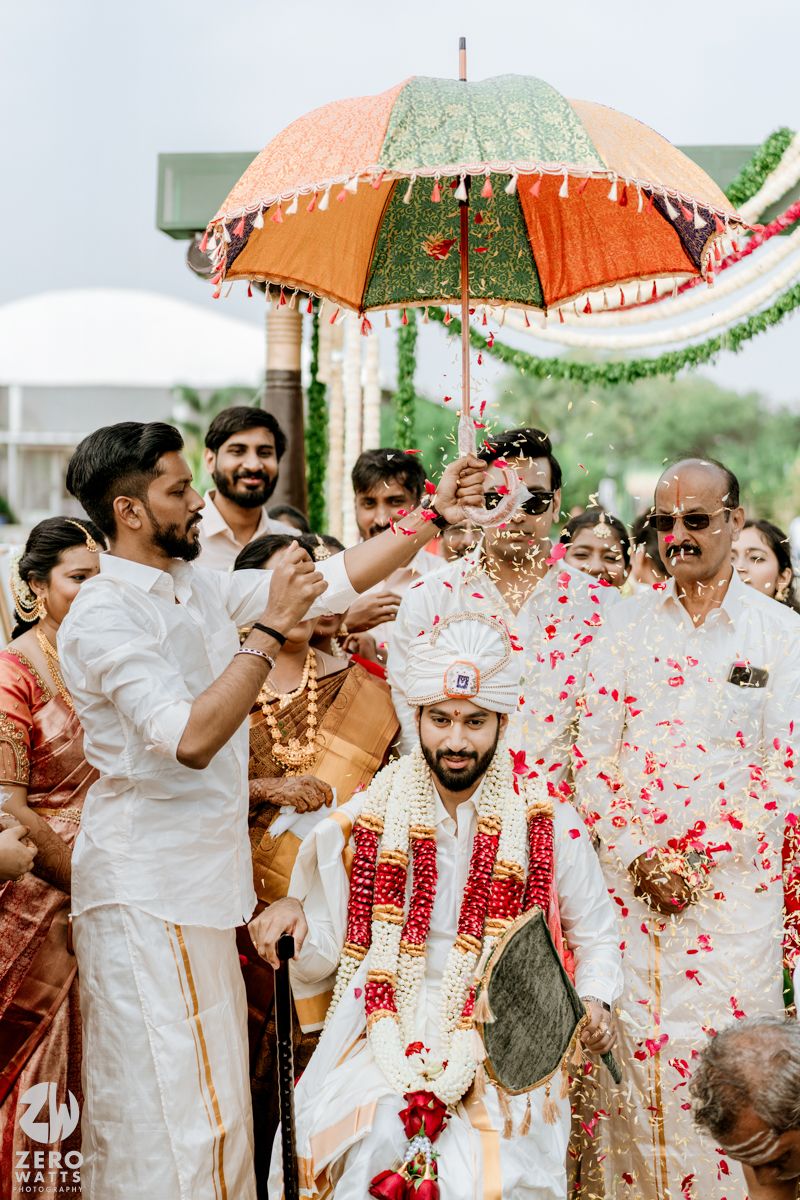 Groom in white sherwani walks under decorative umbrella with flower petals shower at South Indian wedding ceremony.