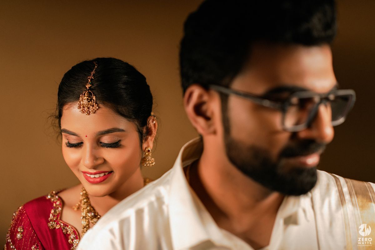South Indian bride in red saree smiling at groom in white shirt during wedding ceremony.