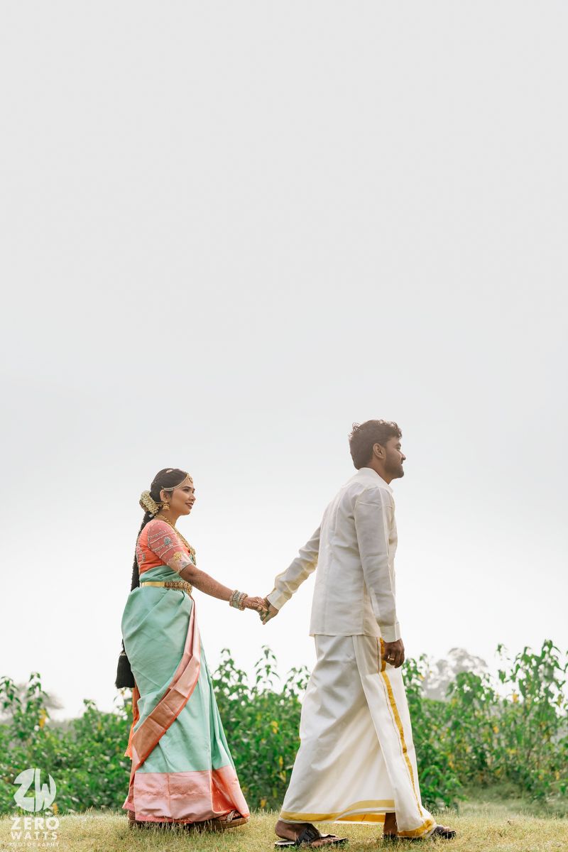 South Indian couple in traditional attire holding hands walking through lush green fields on their wedding day.