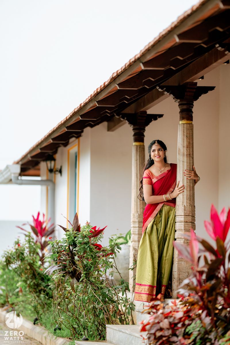 Bride in red and green Kanjivaram saree posing gracefully by a stone pillar of a traditional South Indian home.
