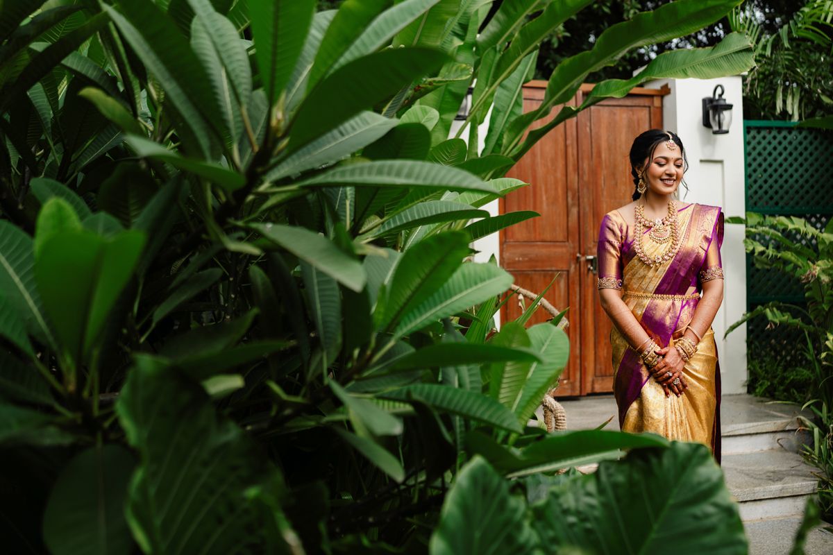A person in a traditional silk saree stands by a wooden door surrounded by lush tropical foliage.