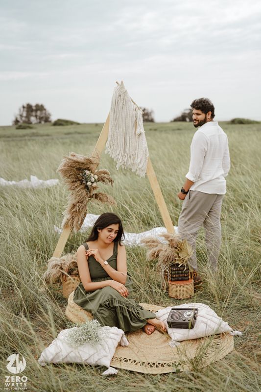 A romantic outdoor picnic setup in a field with white fabric tent, pampas grass decor, and cushions on a round rug.