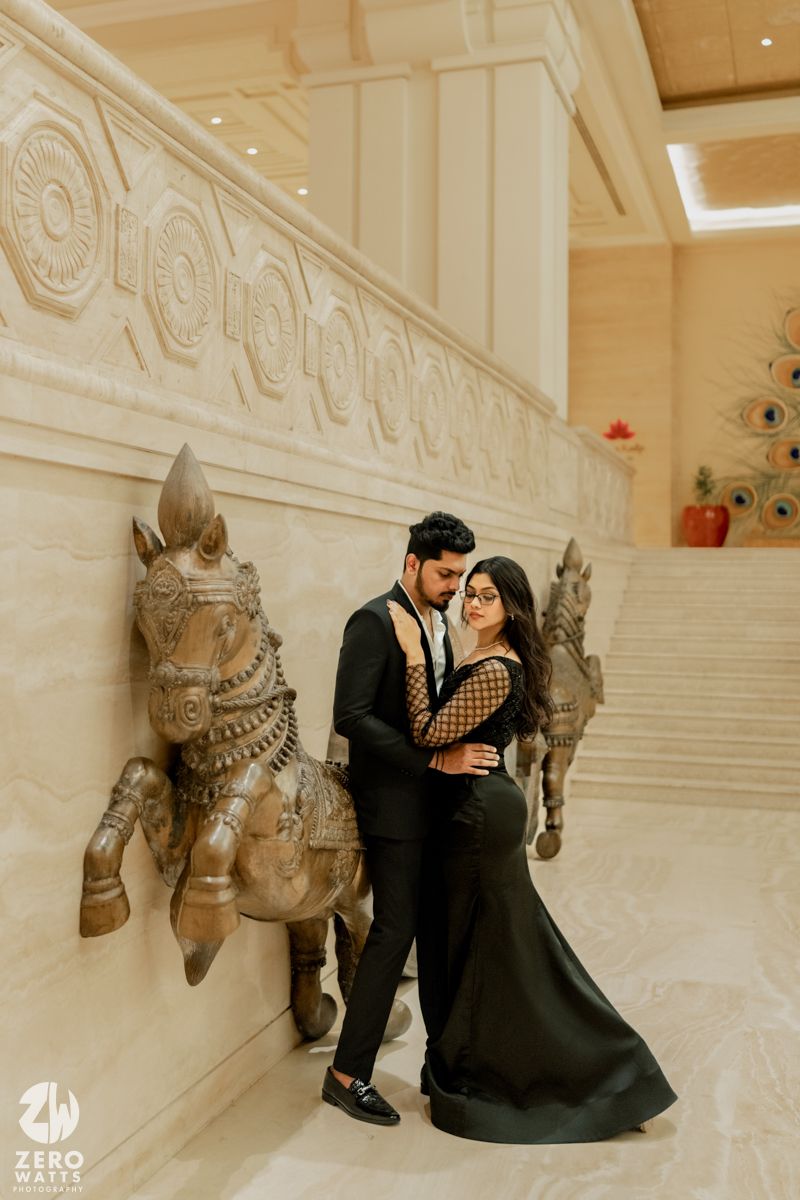 Elegant couple in formal attire pose against ornate white marble wall with decorative elephant sculptures.