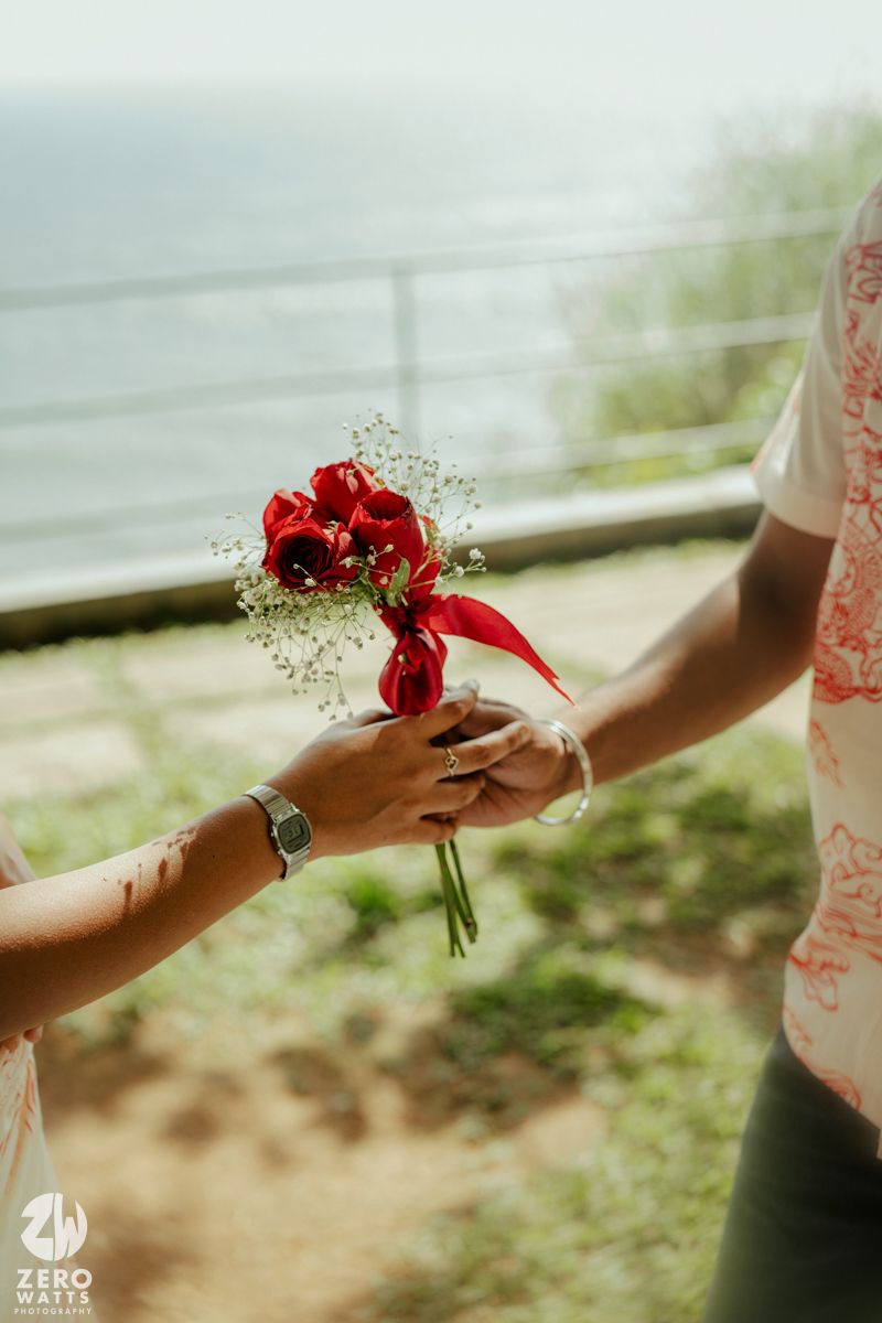 A small red rose bouquet with baby's breath being exchanged between two hands against an ocean backdrop.
