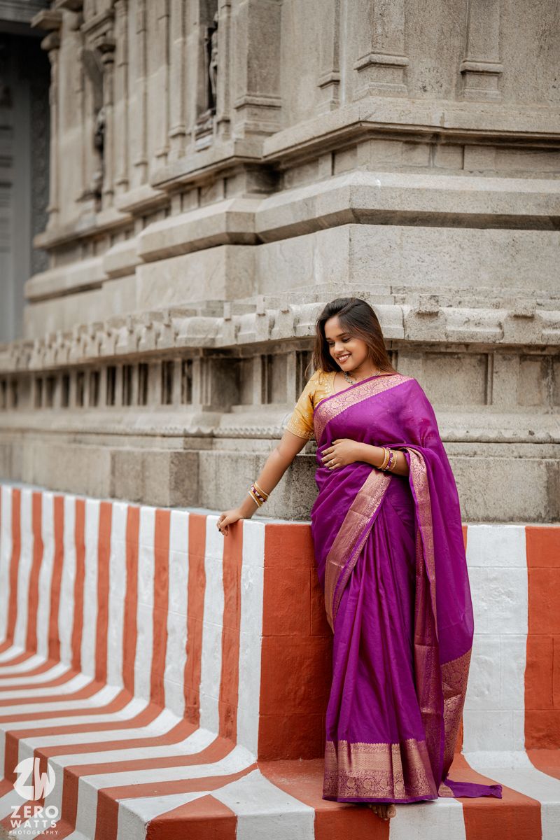 A person in a vibrant purple sari poses against a striped red and white wall at a temple.