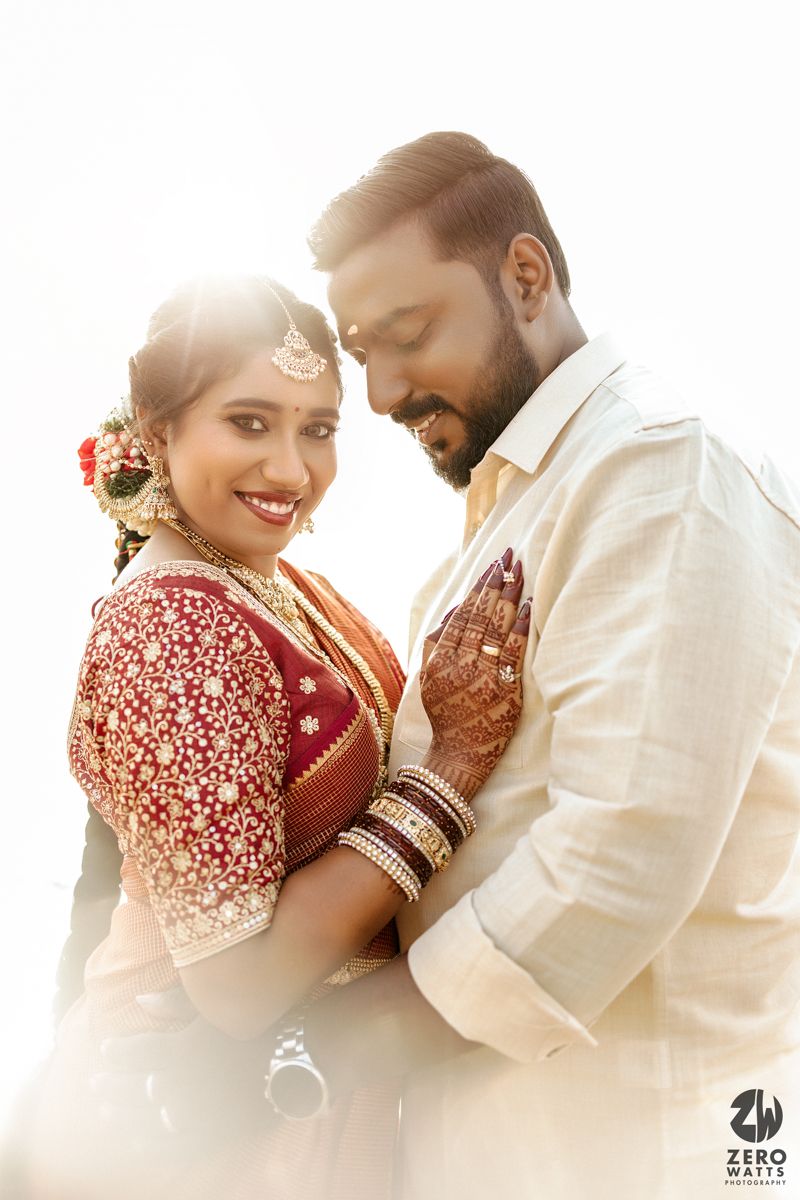 A couple in traditional South Asian wedding attire share an intimate moment in soft sunlight.