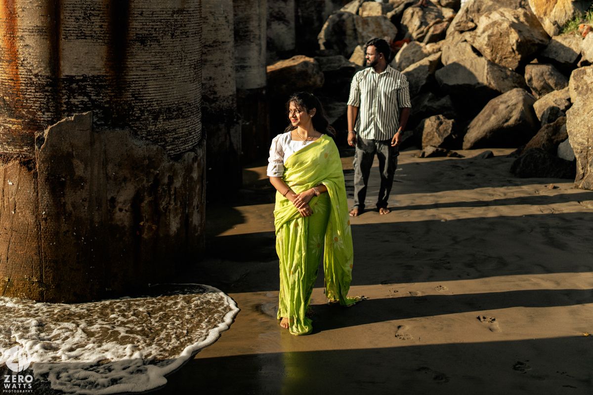 A couple walks along a rocky beach with stone structures in the background.