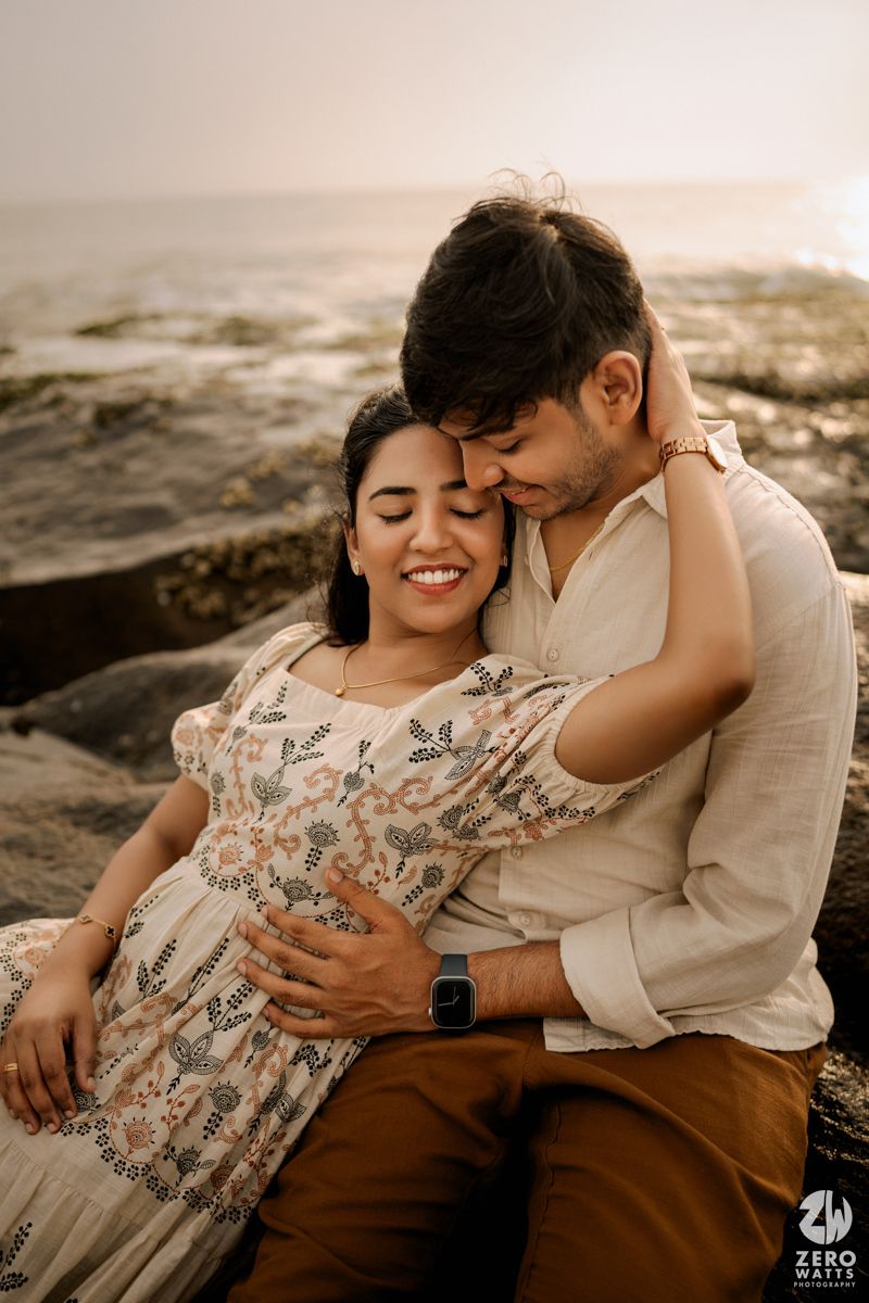 Romantic maternity photoshoot on rocky beach with couple in floral dress and casual attire.