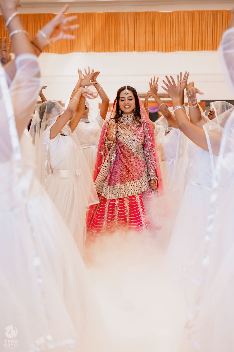 A bride in traditional Indian attire celebrates with guests in white raising their hands during a wedding ceremony.