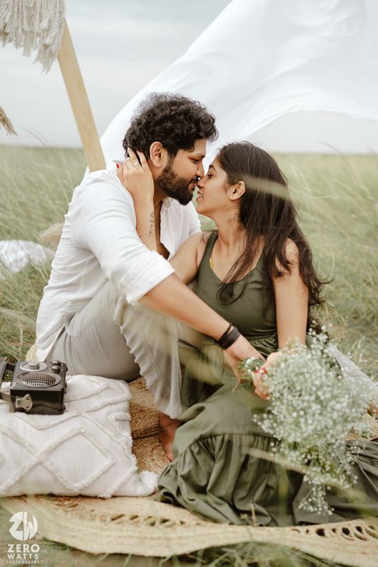 A romantic couple shares tender moments on a picnic blanket surrounded by delicate white flowers in a grassy field.