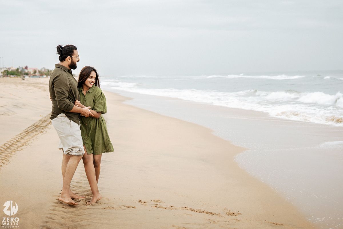 A couple embraces while standing on a peaceful beach with waves and cloudy skies in the background.