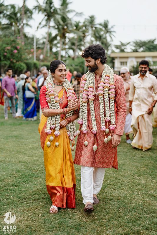 Wedding ceremony on a lawn with palm trees where couple walks together wearing flower garlands.