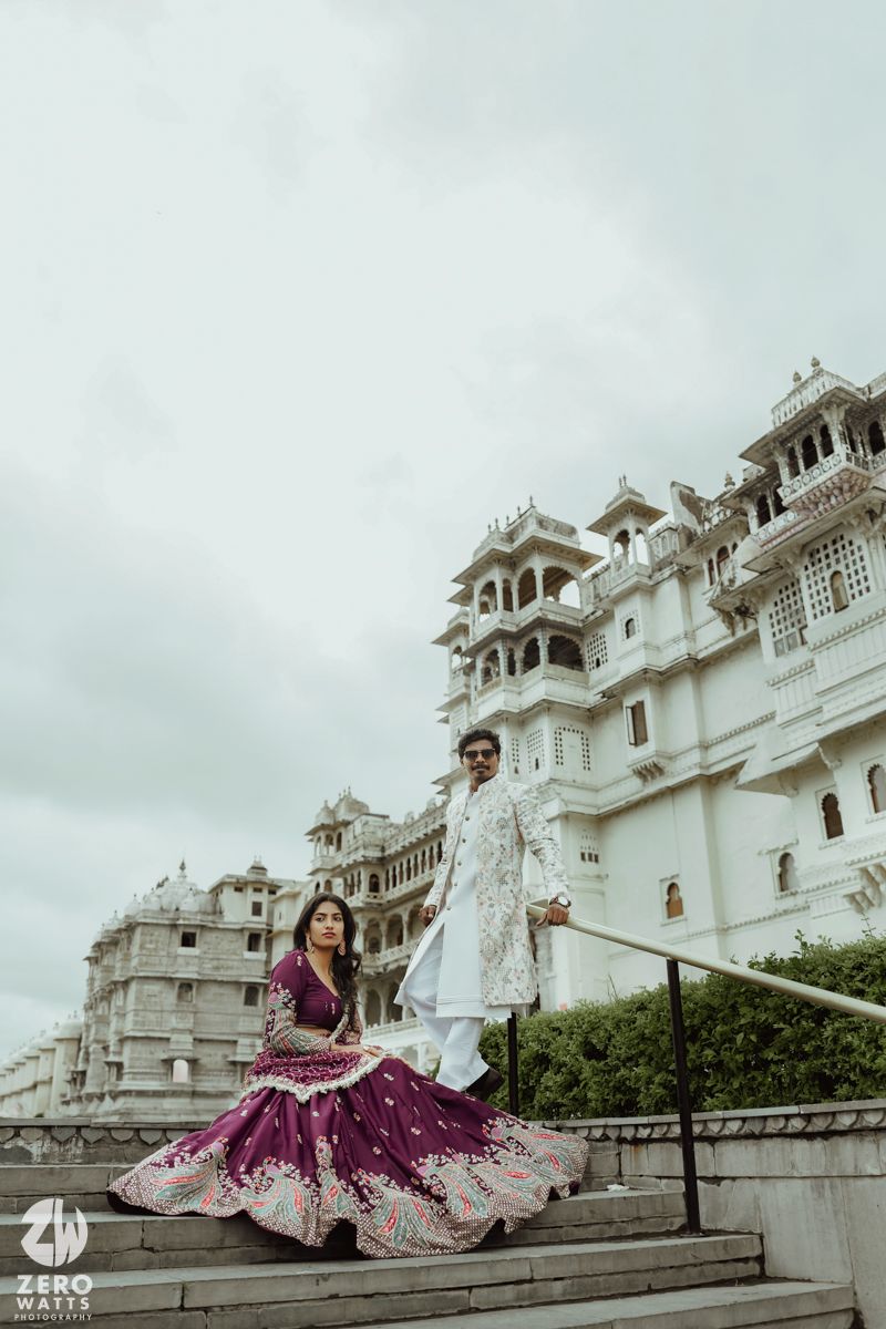 A couple poses against a stunning white palace backdrop in Udaipur with ornate architecture and cloudy skies.