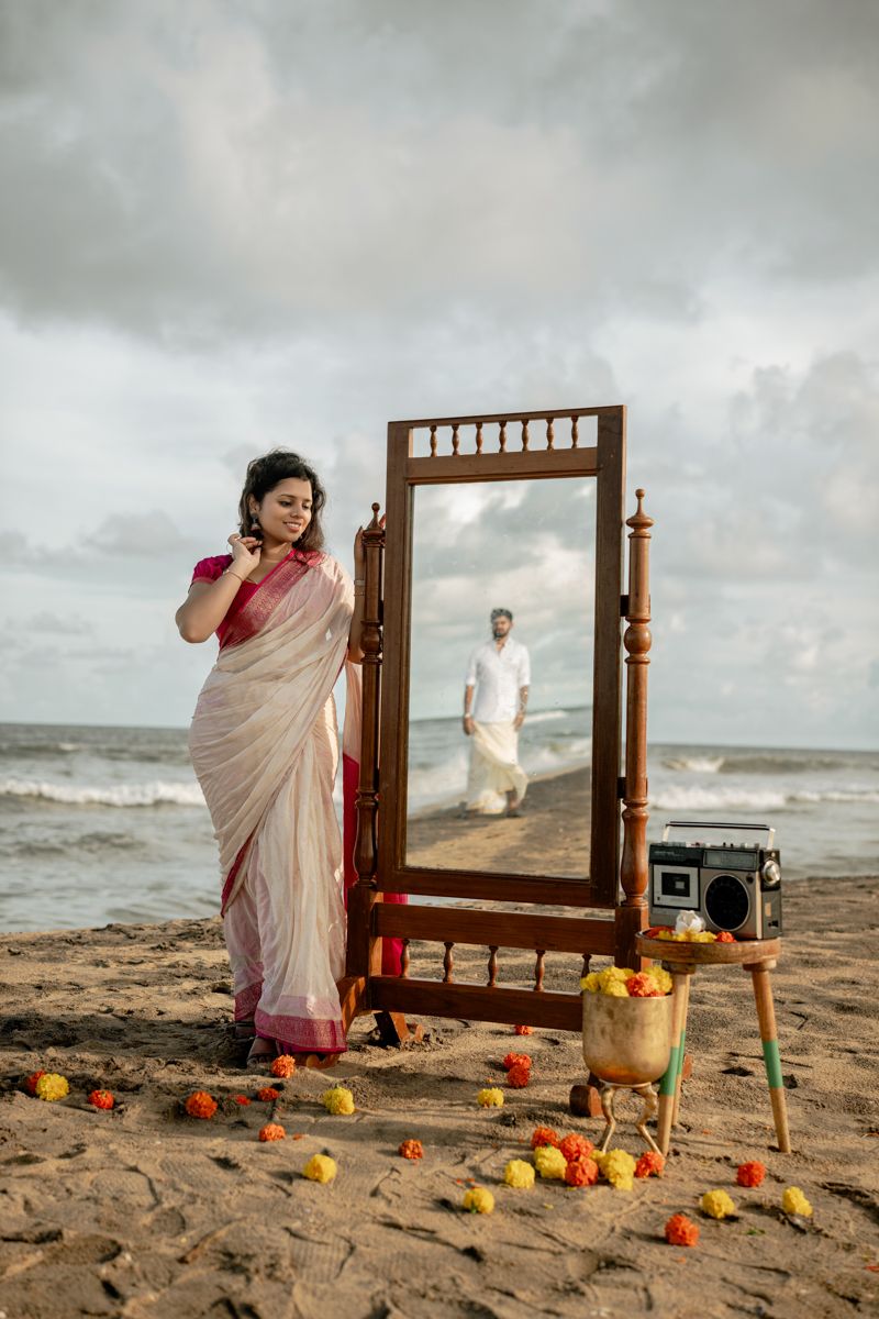 Wooden mirror frame and decorative elements arranged on a beach with scattered orange flowers and crashing waves.