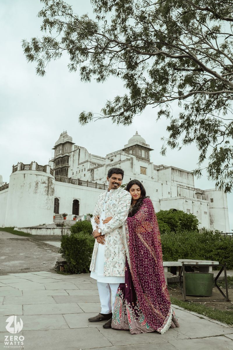 A couple poses against a white palace backdrop wearing traditional Indian wedding attire in purple and white.