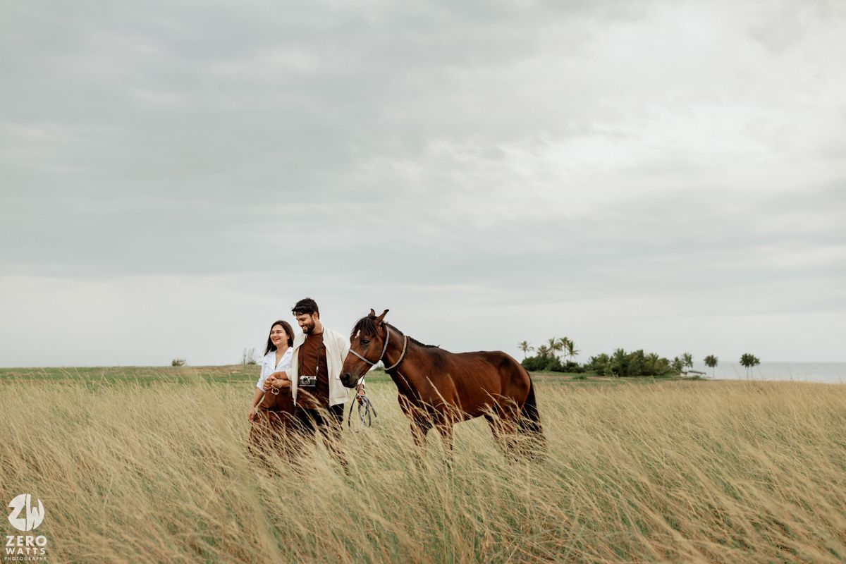 Wide panoramic view of a couple with horses in a windswept prairie field under cloudy skies.