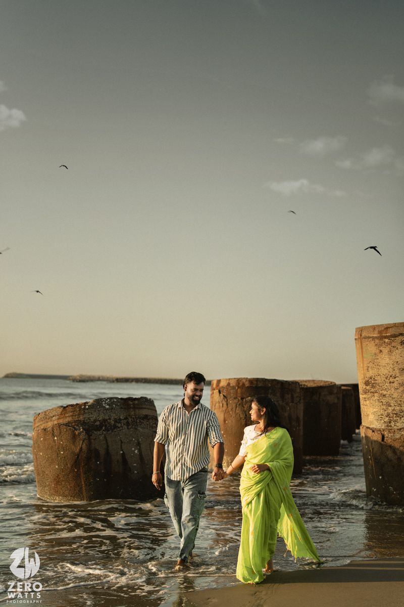 Romantic couple in contrasting outfits pose playfully among concrete pillars at a moody beach during sunset.