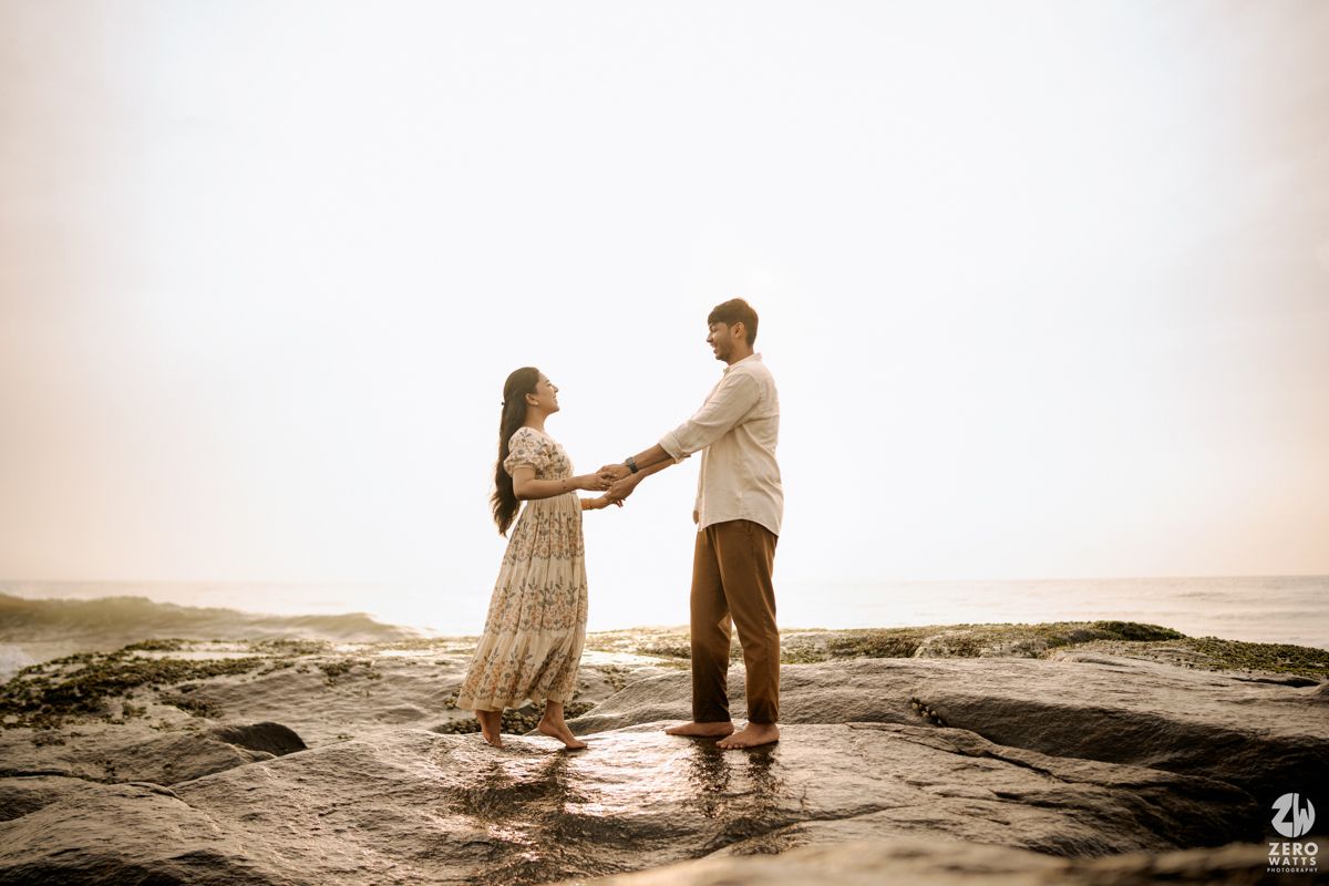 A romantic couple stands on rocky coastal cliffs at sunset creating silhouettes against the golden sky.