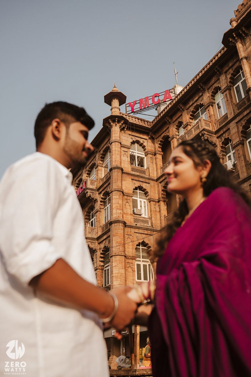 Couple shares a romantic moment against the backdrop of an ornate red sandstone building with intricate architecture.